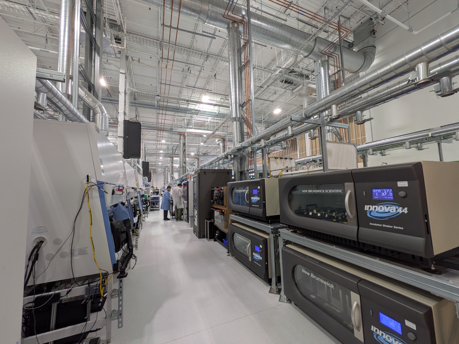 A long aisle of the automated laboratory floor at Emerald Cloud Lab, lined with incubator shakers whose displays show live speed and temperature readings, receding toward scientists working in the distance.
