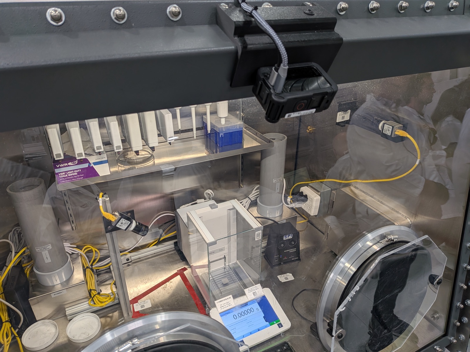 Inside a glovebox at Emerald Cloud Lab: an analytical balance reading 0.00000 g, flanked by two mounted cameras that record every measurement, with QR-code labels on the equipment and work surfaces.
