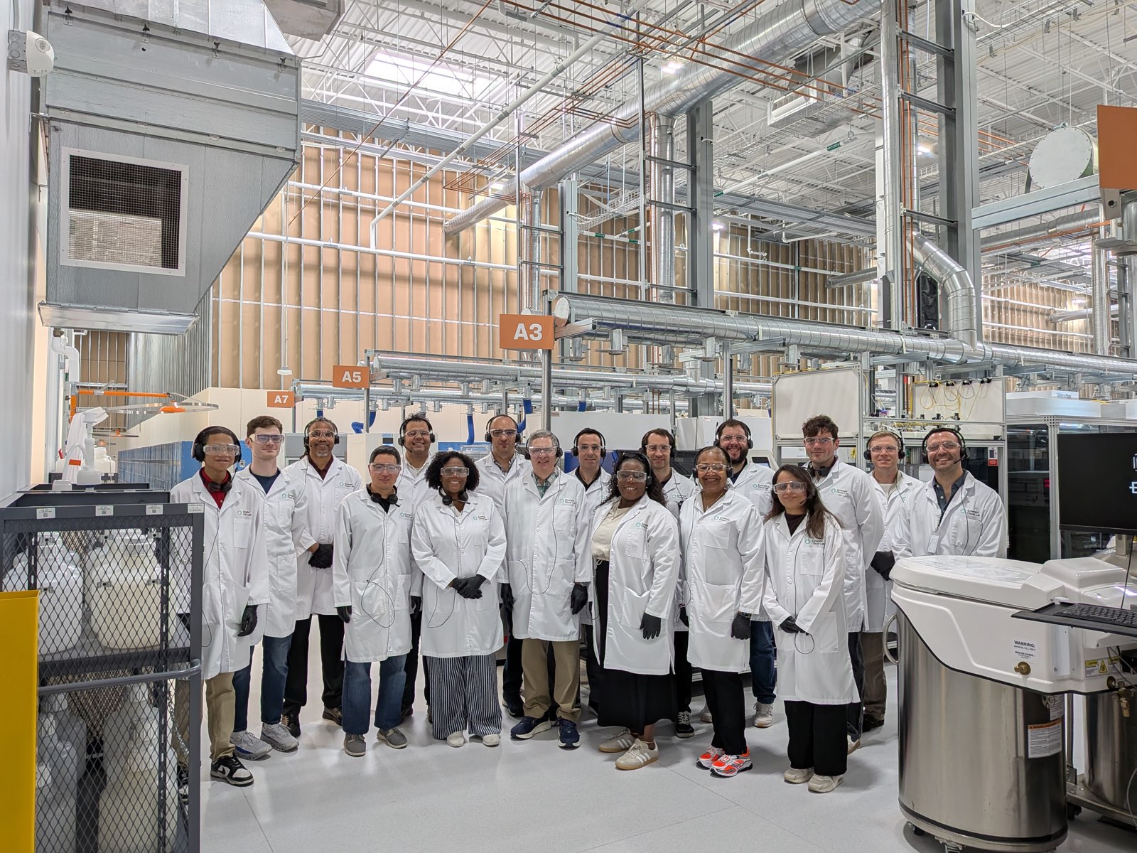 Part of the GEMSTONE team, in lab coats and safety glasses, standing together on the automated laboratory floor at Emerald Cloud Lab in Austin, Texas.