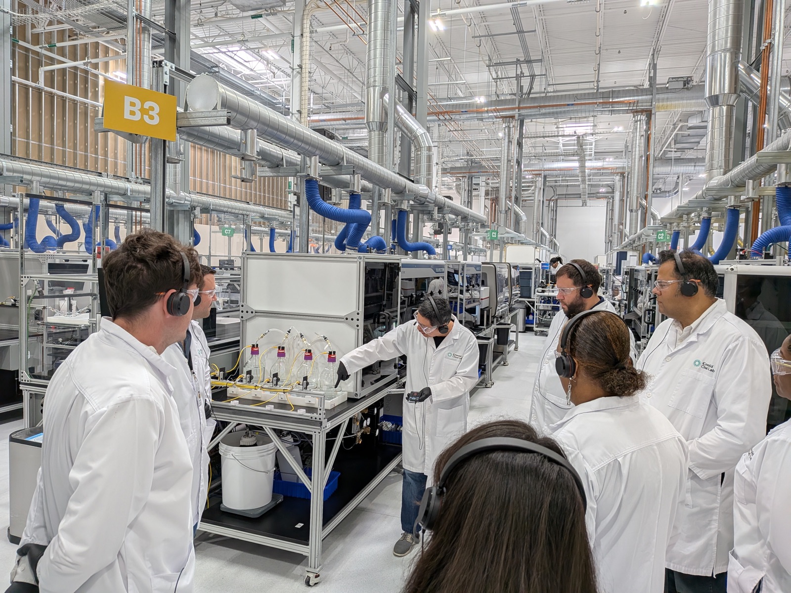 An Emerald Cloud Lab scientist demonstrates a station of sensor-capped reagent bottles to GEMSTONE team members wearing lab coats, safety glasses, and tour headsets on the automated laboratory floor.