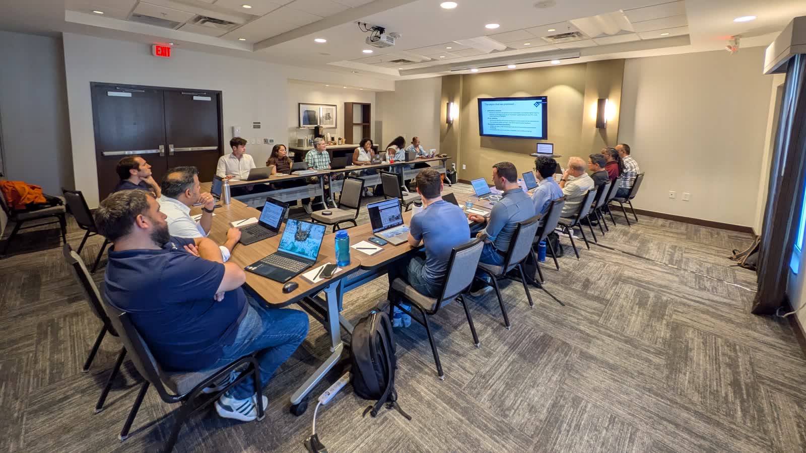 The GEMSTONE team seated around U-shaped tables with laptops during a planning session, facing a screen showing a slide titled 'The vision that we promised'.